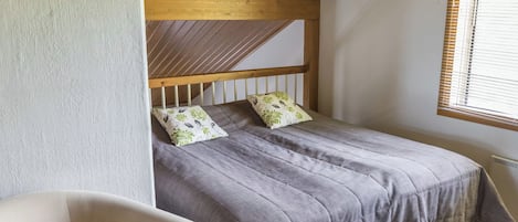 Una camera da letto con soffitto spiovente, un letto con biancheria grigia, due cuscini con motivi floreali, una testiera in legno, una sedia e una finestra con persiane.