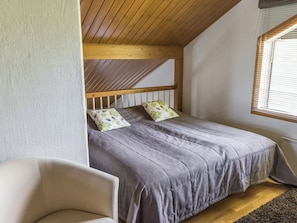 A bedroom with a sloped ceiling, a bed with grey bedding, two pillows with floral patterns, a wooden headboard, a chair, and a window with blinds. - Alppinuppu by Interhome (Nilsiä)