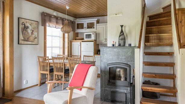 A cozy living room with a fireplace, wooden dining table, and chairs. A staircase leads to an upper level. - Alppinuppu by Interhome (Nilsiä)