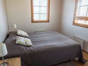 Un dormitorio con una cama, dos almohadas, una lámpara de noche, un radiador y una ventana con persianas.