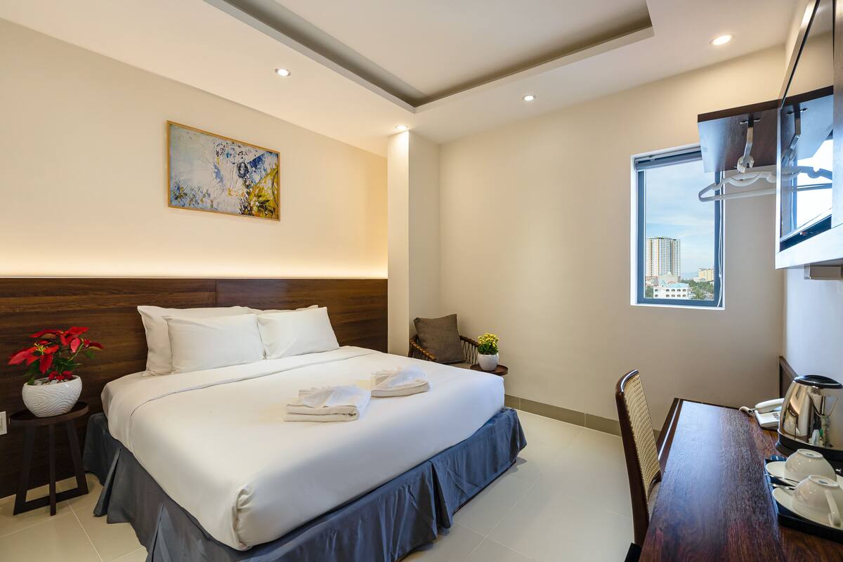 A modern hotel room with a large bed, a small table with a kettle, a chair, and a window with a city view.