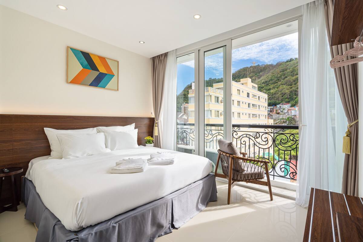 A hotel room with a large bed, a view of buildings through a balcony, and a decorative wall piece.