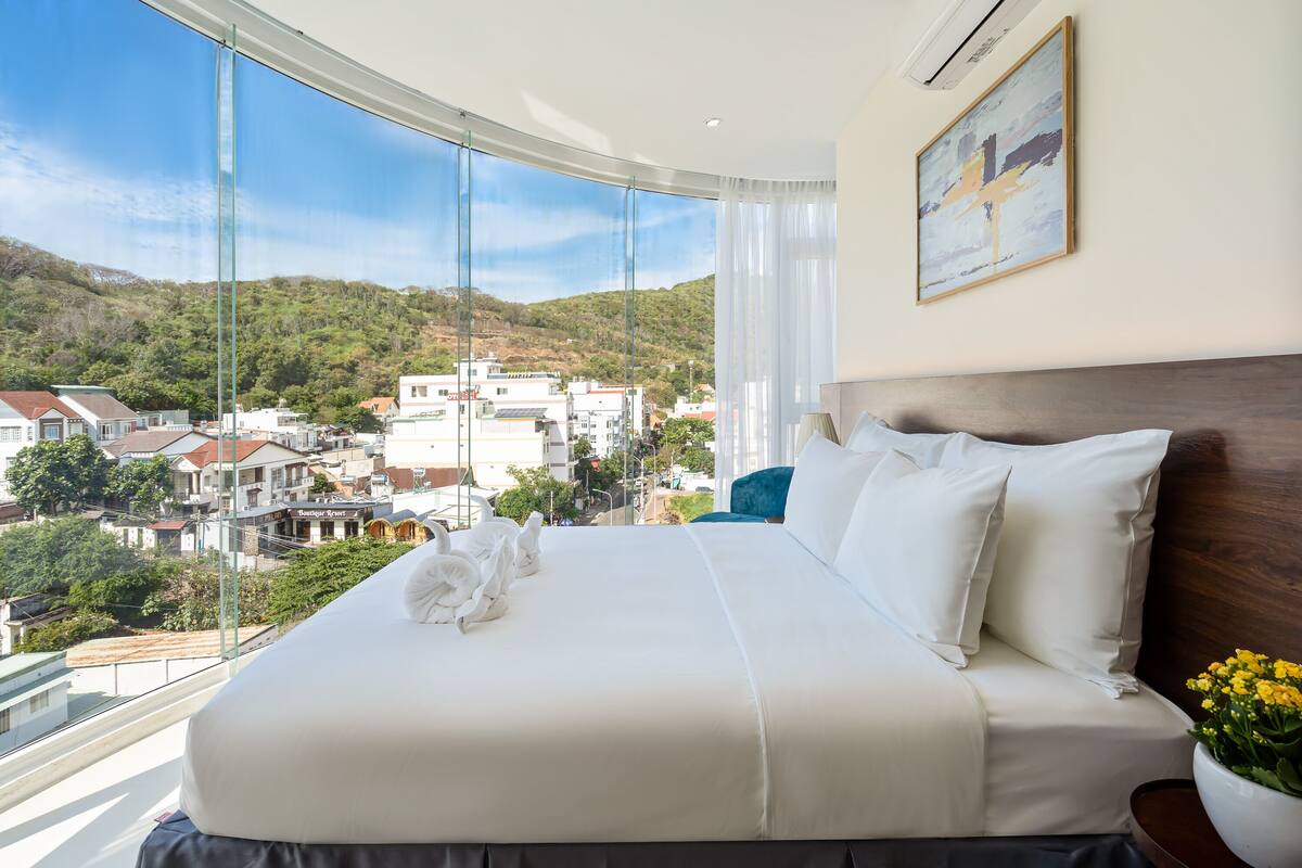 A hotel room with a large bed, white bedding, a view of a town and hills, and a painting on the wall.