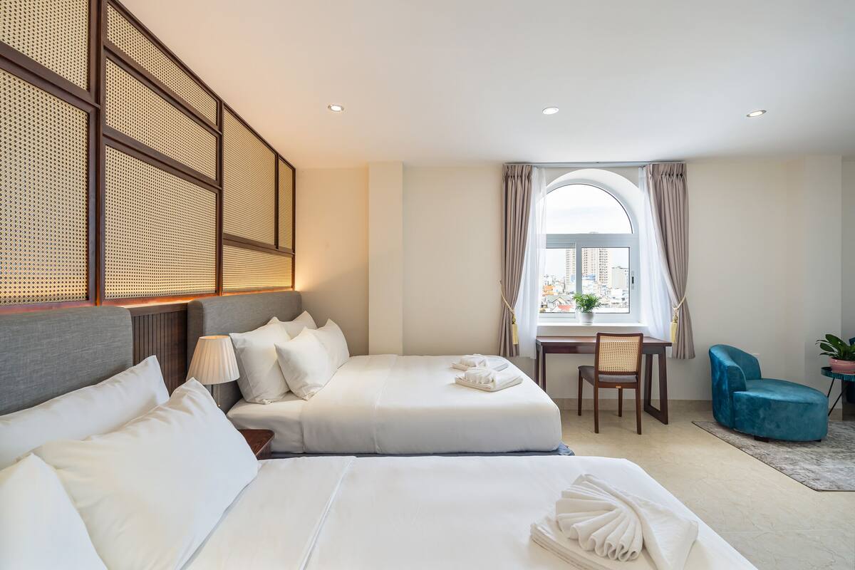 A hotel room with two beds, a desk, a chair, and a view of the city through a large window.