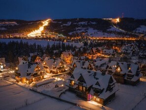 Un pueblo cubierto de nieve con edificios de estilo chalete tradicional, un telesilla al fondo y una zona de estación de esquí iluminada.