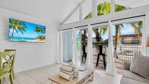 A spacious room with a large TV displaying a beach scene, a wooden coffee table with towels, and a view of the outdoor patio with palm trees. - Beach Condo with Pool Near Holmes Beach! (Holmes Beach)