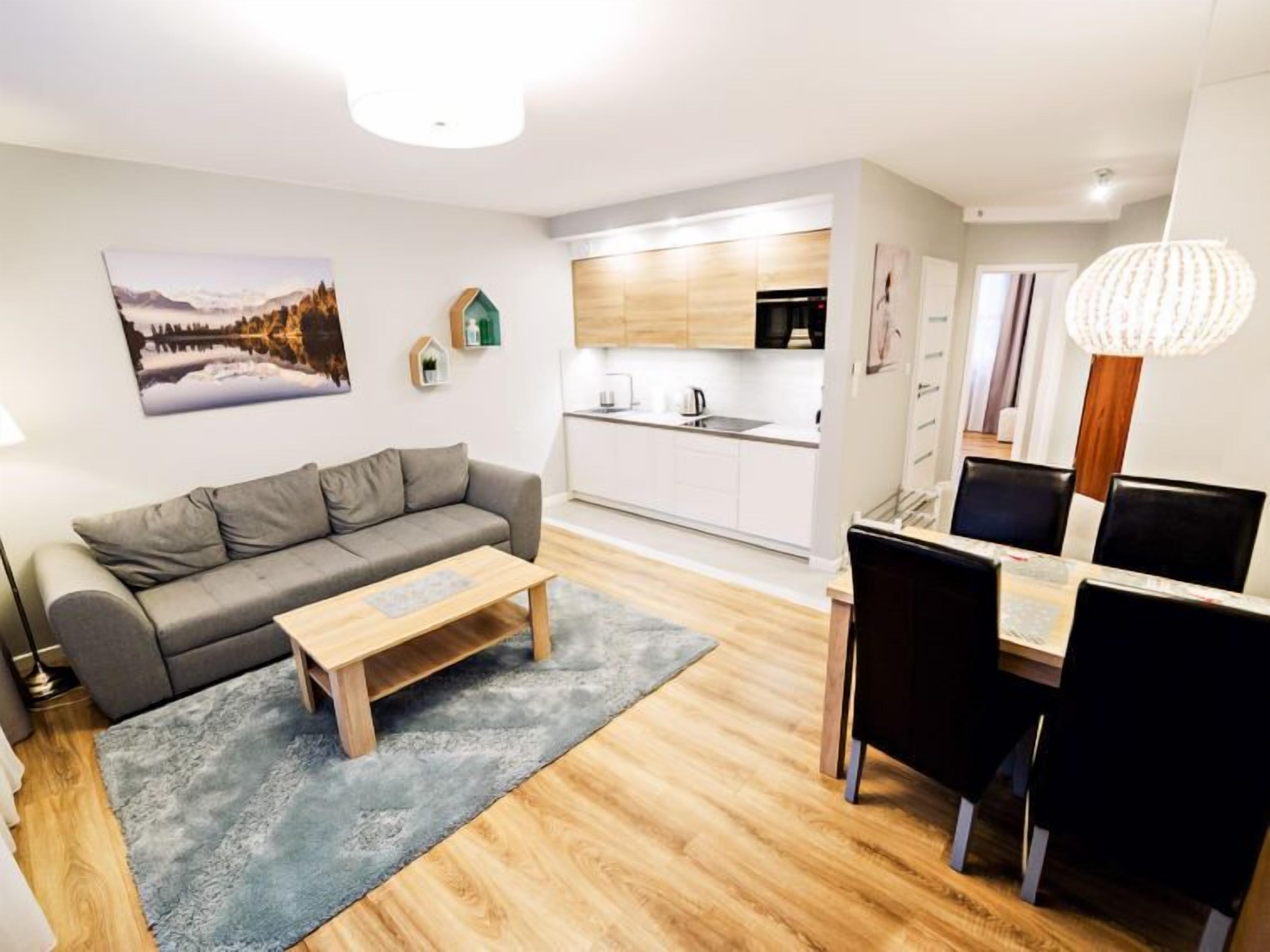Una sala moderna con un sofá gris, una mesa de centro de madera y un comedor con una mesa y sillas negras. Se ve una cocina con gabinetes blancos y una encimera de madera al fondo.