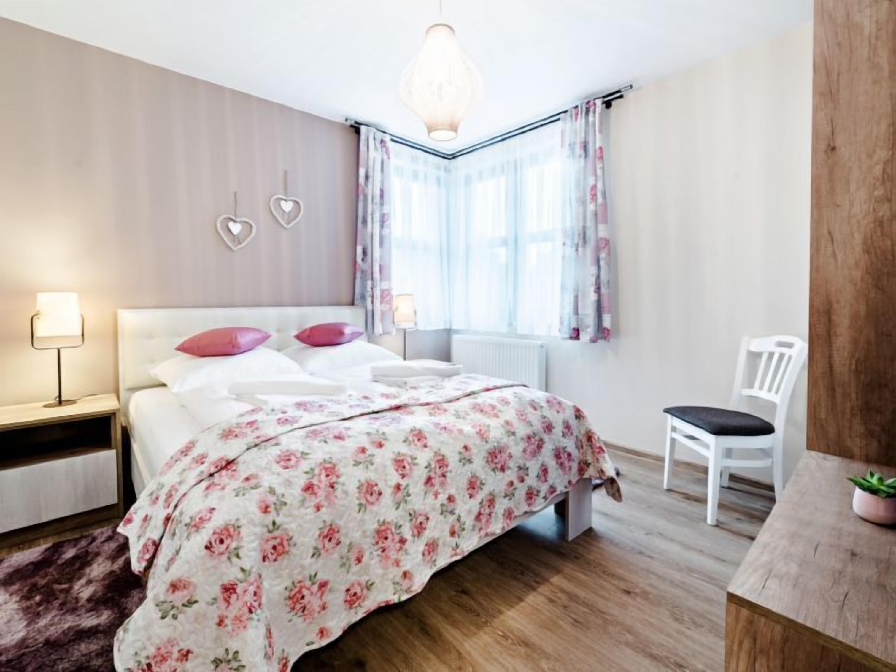 Un dormitorio con cama, lámparas de noche, una silla, una ventana con cortinas y una lámpara colgante.