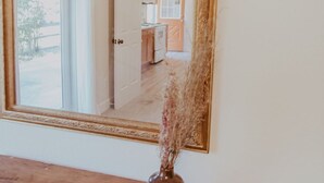A large mirror with a decorative frame on a wooden surface. In the reflection, a doorway leads to a room with a kitchen and a view of a beach. - Tadpole Cottage (Cloverdale)