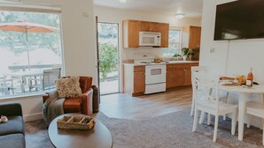 A modern kitchen with wooden cabinets and a white microwave, a dining area with white chairs, and a cozy seating area with a brown sofa and a small table. - Tadpole Cottage (Cloverdale)