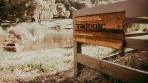 A wooden sign with the name 'Tadpole' in a rustic font, mounted on a wooden fence with a natural landscape in the background. - Tadpole Cottage (Cloverdale)