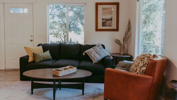 A living room with a dark sofa, a round coffee table, and a brown armchair. There is a framed picture on the wall and a window with a view of trees. - Tadpole Cottage (Cloverdale)