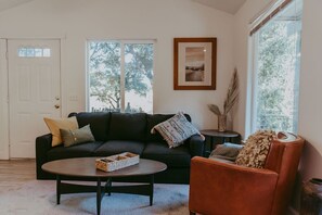 A living room with a dark sofa, a round coffee table, and a brown armchair. There is a framed picture on the wall and a window with a view of trees. - Tadpole Cottage (Cloverdale)