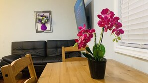 Una sala de estar con una mesa de comedor de madera, un sofá de cuero negro, una silla de madera y una planta de orquídea en maceta.