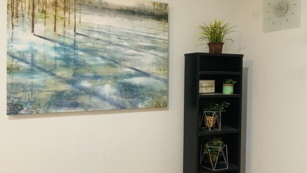 Una habitación con un cuadro abstracto grande en la pared, un estante negro con plantas en macetas y un reloj de pared.