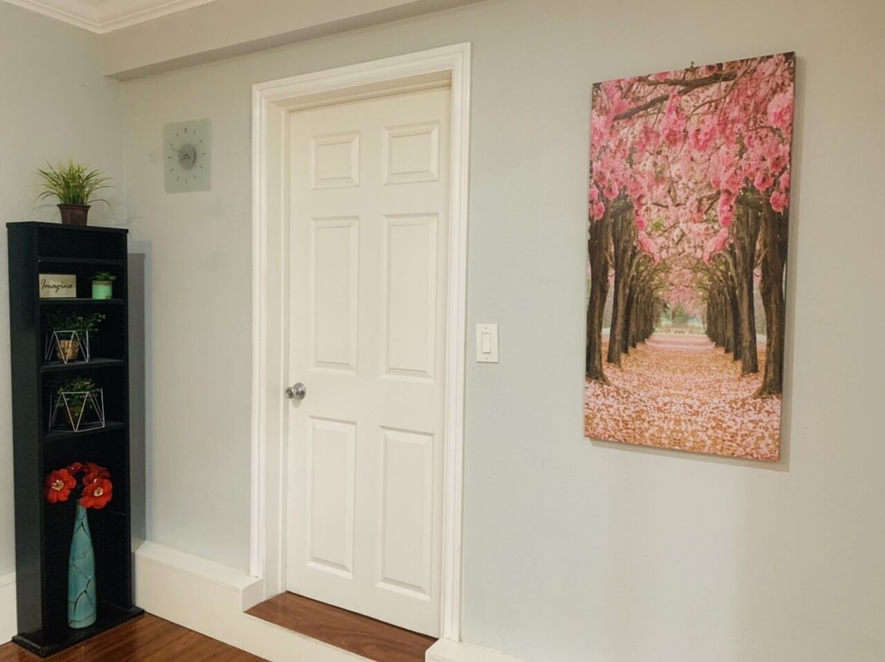 A hallway with a white door, a black shelving unit with decorative items, and a painting of a cherry blossom path.