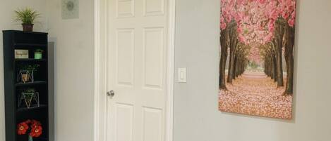 A hallway with a white door, a black shelving unit with decorative items, and a painting of a cherry blossom path.