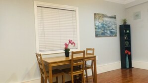 Un comedor con una mesa y sillas de madera, una ventana con persianas, un cuadro en la pared y una alta estantería negra con flores rojas.