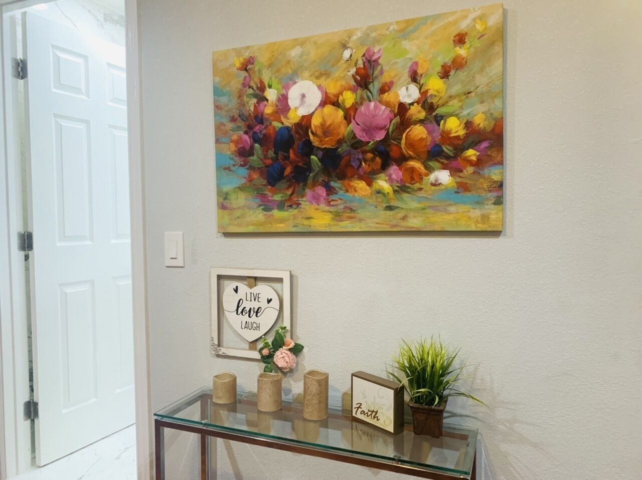 Une pièce avec une console en verre, un cadre contenant une citation inspirante, une plante en pot et une peinture florale accrochée au mur.
