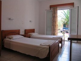 Habitación con dos camas individuales, una mesita de noche de madera, una pequeña mesa y un ventanal corredero que da a un balcón con vistas a un coche aparcado y follaje.