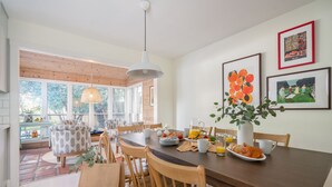 Uma sala de jantar com mesa de madeira, cadeiras e um vaso com flores. Há quadros nas paredes e uma janela com vista para o verde.