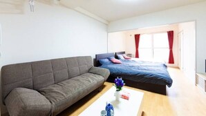 Une chambre moderne avec un canapé gris, un lit avec une literie bleue et une table basse blanche agrémentée d’un vase et d’une boîte.