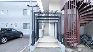 Une entrée fermée par un portail, avec des marches menant à un bâtiment, une voiture garée et des vélos.