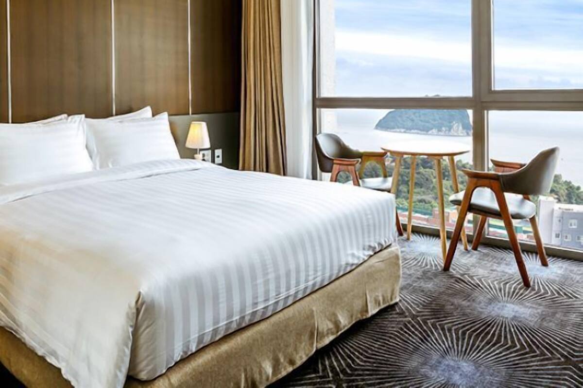 A hotel room with a large bed, a desk with a chair, and a view of the city through large windows.