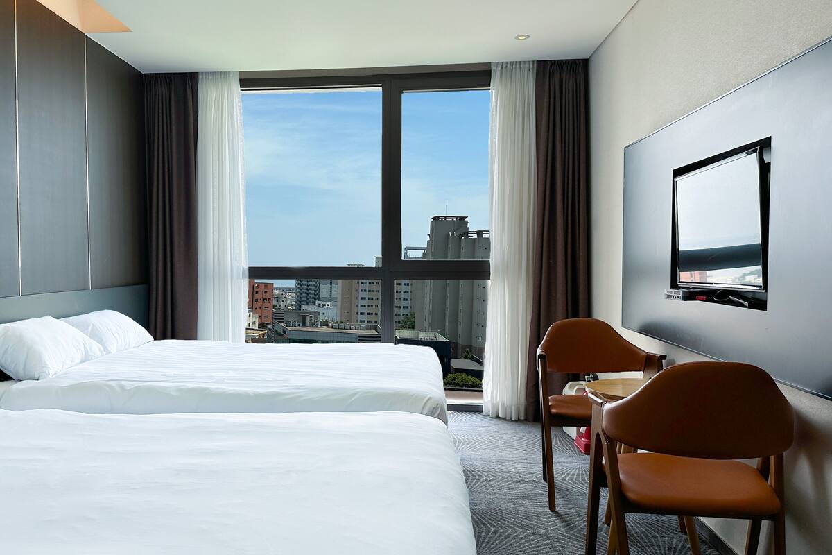 A hotel room with two beds, a desk, a chair, a TV, and a large window with a city view.