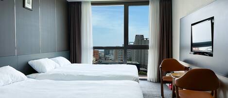 Habitación de hotel con dos camas, un escritorio, una silla, un televisor y un ventanal grande con vistas a la ciudad.