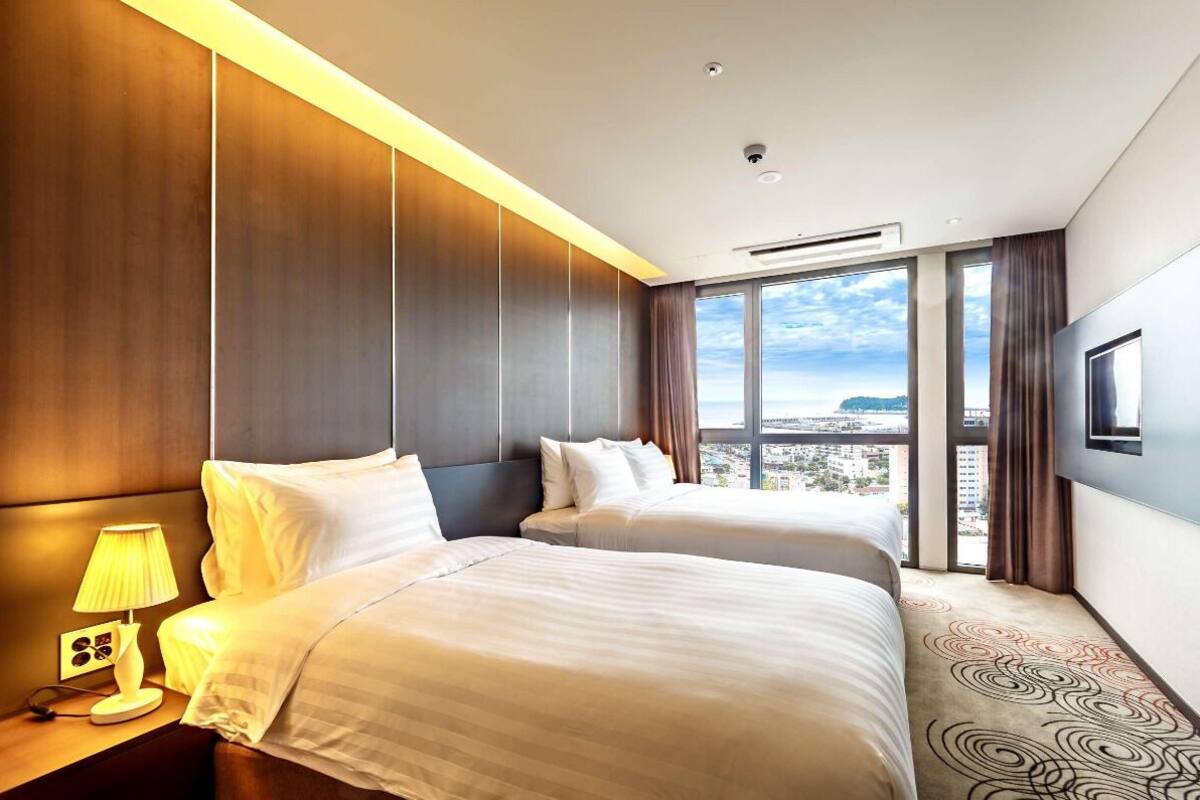 A hotel room with two beds, a large window with a city view, a flat-screen TV mounted on the wall, and a patterned carpet.
