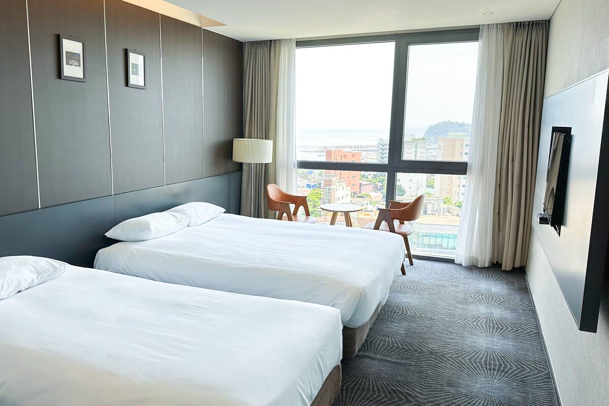 A hotel room with two beds, a small table, two chairs, a lamp, and a large window with a view of buildings.