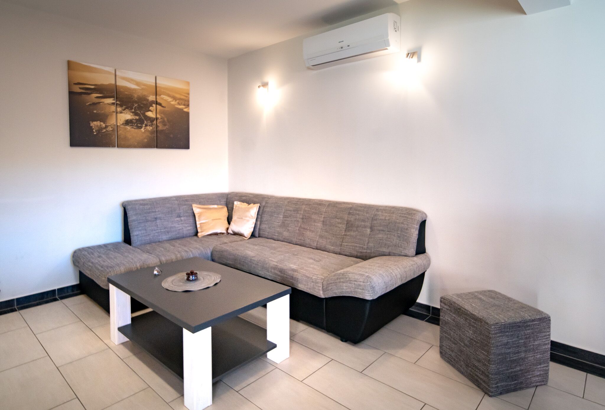 A modern living room with a grey sectional sofa, a dark coffee table, and a small ottoman. There is a wall-mounted air conditioner and a framed picture above the sofa.