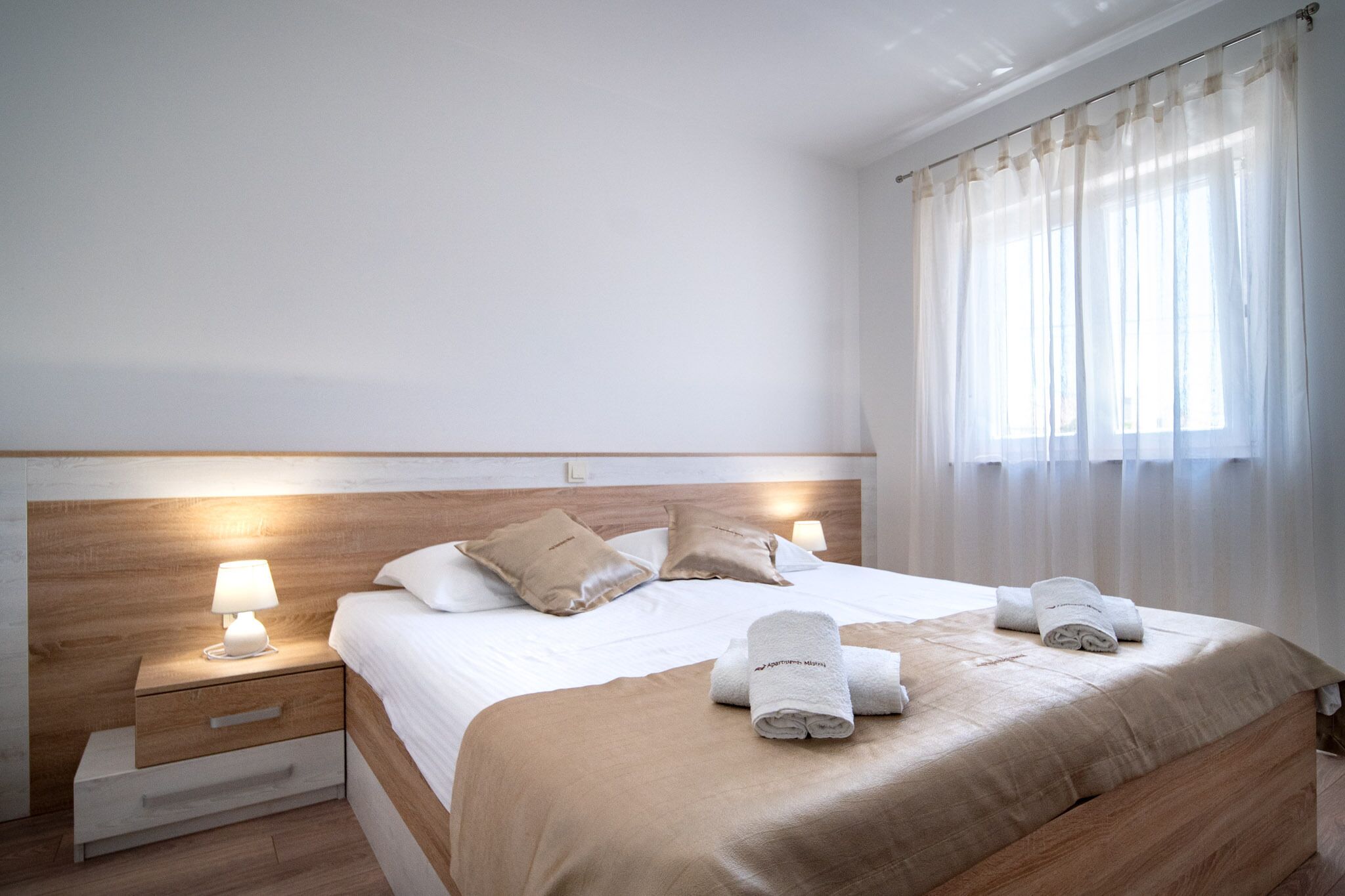 A neatly made bed with a wooden headboard, two bedside lamps, and a window with sheer curtains.