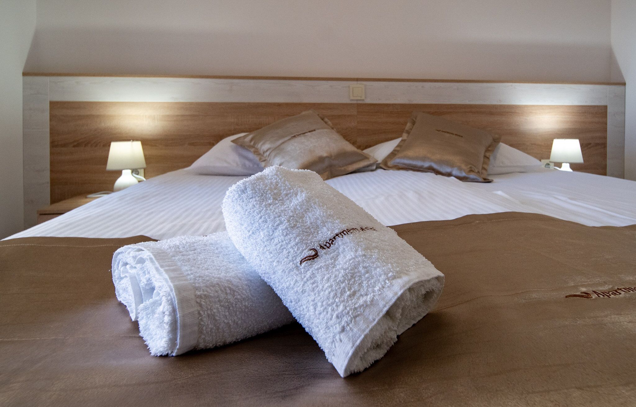 Un lit bien fait, avec du linge de lit blanc et deux serviettes roulées ornées de motifs brodés, une tête de lit en bois et des lampes de chevet.