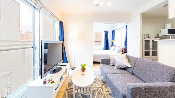 A modern hotel room with a large window, a flat-screen TV, a sofa, a small table, and a kitchen area. - Eve (Sapporo)