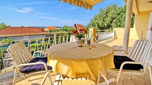 A round table with a yellow tablecloth, surrounded by white metal chairs with cushions, set on a balcony with a view of houses and trees. - Apartment Pehar (Orebic)