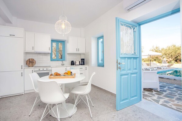 Una cocina moderna con armarios blancos, una mesa de comedor con sillas y una puerta azul que da a una zona de piscina.