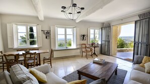 A spacious living room with a large sofa, a coffee table, and a dining area with a table and chairs. There are large windows offering a view of the outdoors. - Séguret, cosy village house with exceptional view (Seguret)