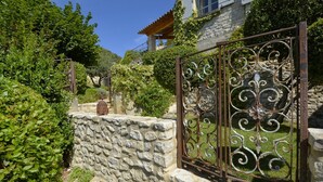 A stone wall with a decorative metal gate, lush greenery, and a building with a wooden roof in the background. - Séguret, cosy village house with exceptional view (Seguret)