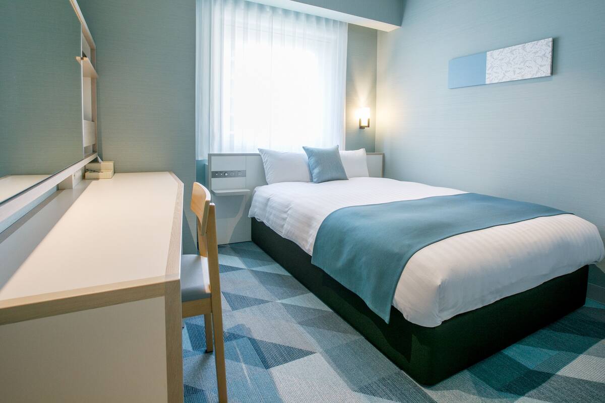Une chambre d’hôtel comprenant un lit, un bureau, une chaise, une table de chevet, une lampe et une fenêtre avec des rideaux.