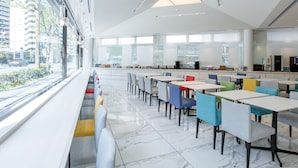 Uma cafeteria moderna com pisos de mármore branco, cadeiras coloridas e um grande janelão com vista para a cidade.