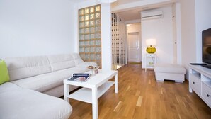 Una sala de estar moderna con un sofá blanco, una mesa de centro blanca y un televisor sobre un mueble blanco. Hay una lámpara amarilla sobre una mesita, y un armario blanco con cajones.