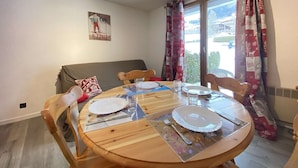 A dining area with a round wooden table set for four, a grey sofa, and a window with red and white curtains. - Cozy Garden-Level Studio Cabin in Praz-sur-Arly - Sleeps 4 (Praz-sur-Arly)