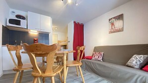 A compact living space with a kitchenette, a dining area with wooden chairs, a sofa with cushions, and a sports-themed wall decoration. - Cozy Garden-Level Studio Cabin in Praz-sur-Arly - Sleeps 4 (Praz-sur-Arly)