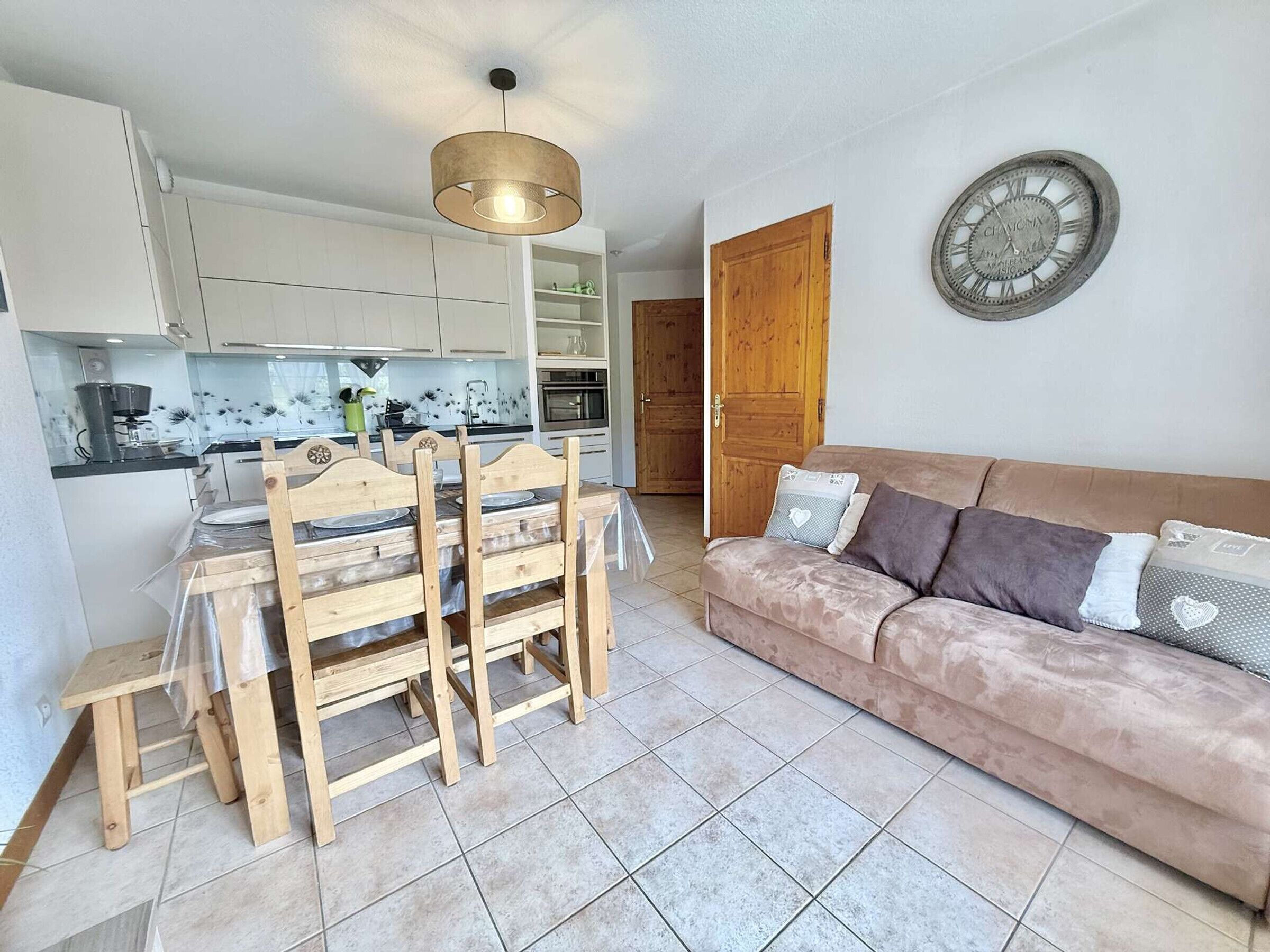 A compact living space with a kitchenette, a dining area with wooden chairs, a cushioned sofa, and a wall clock.