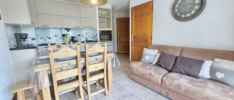A compact living space with a kitchenette, a dining area with wooden chairs, a cushioned sofa, and a wall clock.
