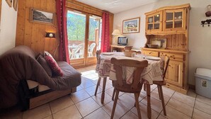 Et hyggeligt værelse med sofa, spisebord med stole, fjernsyn, træskabe og et stort vindue med gardiner.