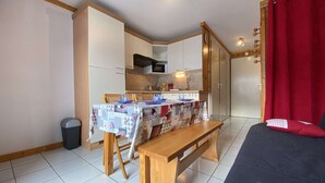 A compact room with a kitchenette, a dining table with a quilted tablecloth, a bench, and a bed with red curtains. - Studio cabin, pets welcome - 4 guests, views of ski slopes (Praz-sur-Arly)
