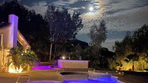 A nighttime outdoor pool area with a moonlit sky, a building with a white facade, and a small waterfall feature.